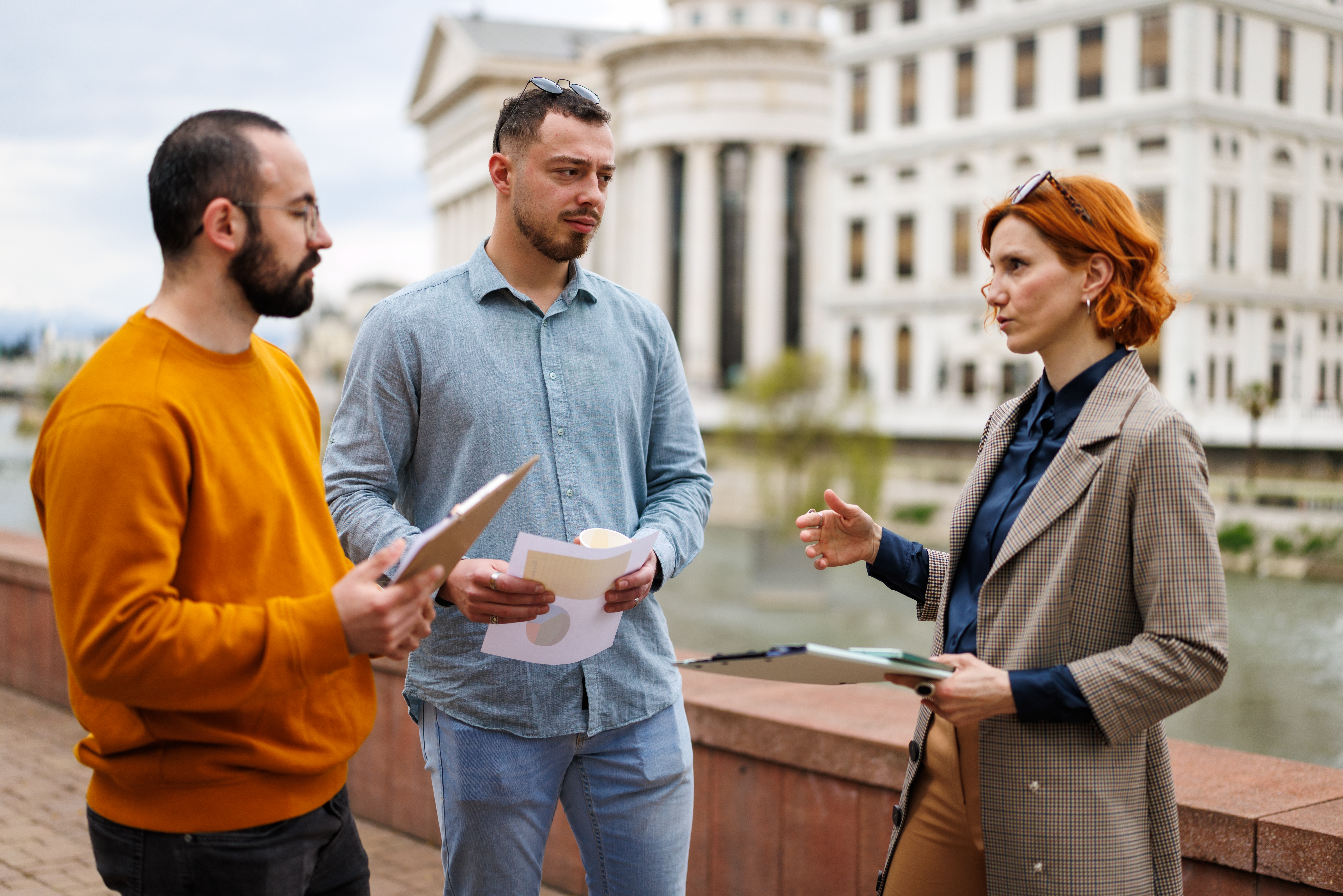 Three individuals engage in conversation near a river. They hold papers and clipboards while discussing various topics as tall buildings rise behind them.